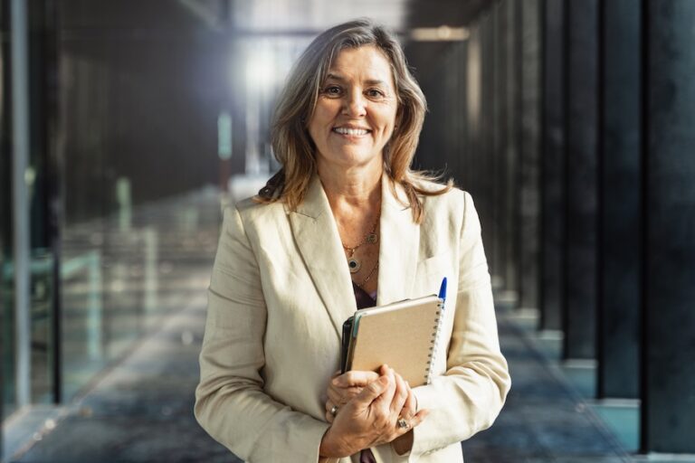 Business woman in a beige blazer holds notebook with a pen in the spine while looking at the camera